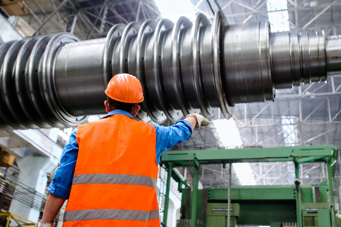 Fachkraft mit Helm und Warnweste prüft eine große Maschinenwelle in einer Industriehalle im Mittelstand
