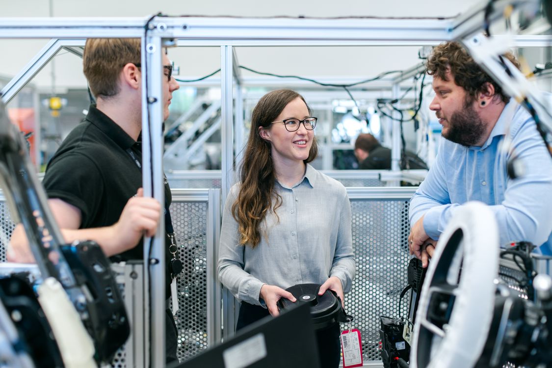 Drei Führungskräfte im Gespräch an einer Montagezelle mit Lenkrädern und Cockpitbauteilen bei einem Automobilzulieferer