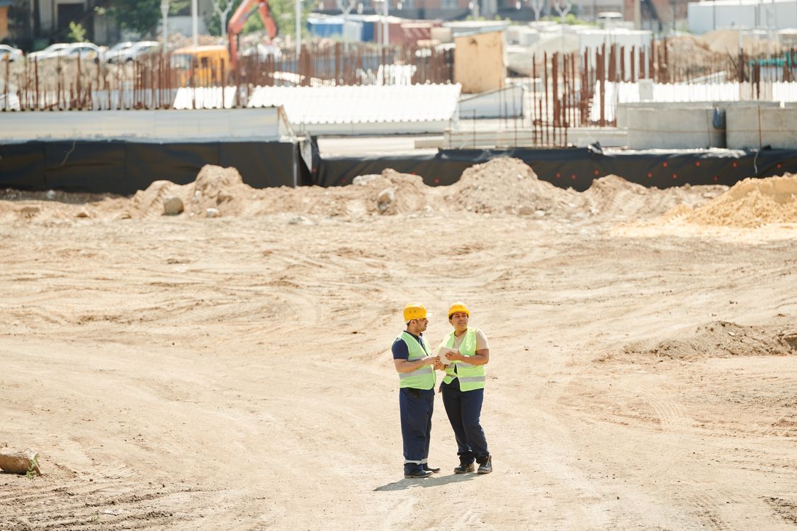 Zwei Bauleiter mit Helm und Warnweste besprechen auf einer großen Baustelle die nächsten Bauabschnitte