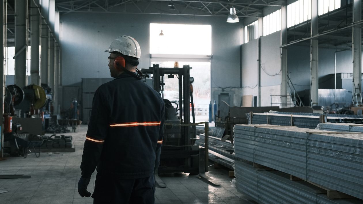 Technische Führungskraft mit Schutzhelm und Gehörschutz überblickt eine Industriehalle mit Maschinen, Stapler und Material
