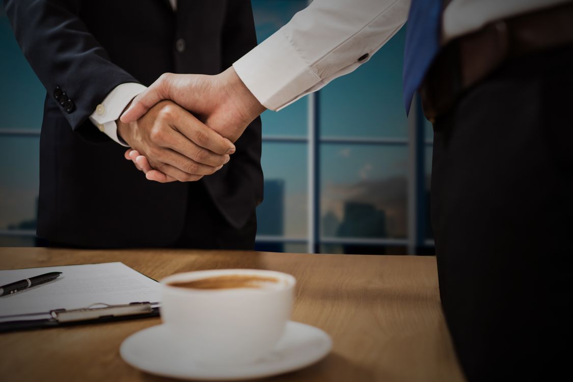 Handschlag zweier Geschäftsleute am Tisch mit Vertragsunterlagen und einer Tasse Kaffee