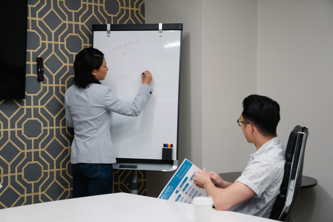 Vertriebsmitarbeiterin schreibt Notizen auf ein Whiteboard, ein Kollege verfolgt die Präsentation